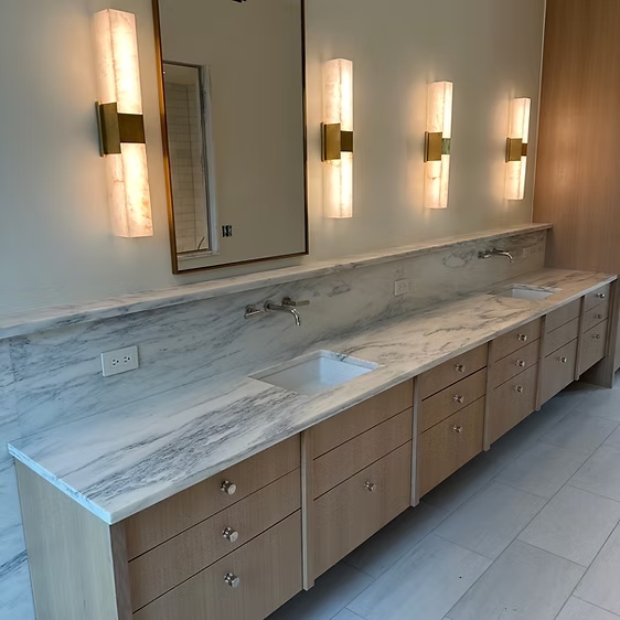 Long double vanity with marble countertop and backsplash, brass wall sconces, and wood cabinetry — a real Precision Granite & Marble project.