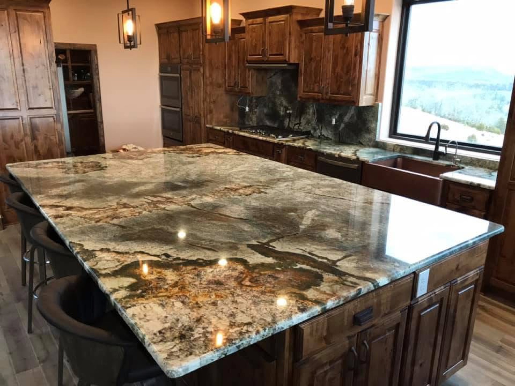 Kitchen island in dramatic gold and green granite with a copper farmhouse sink and Ozark mountain view — a real Precision Granite & Marble installation.